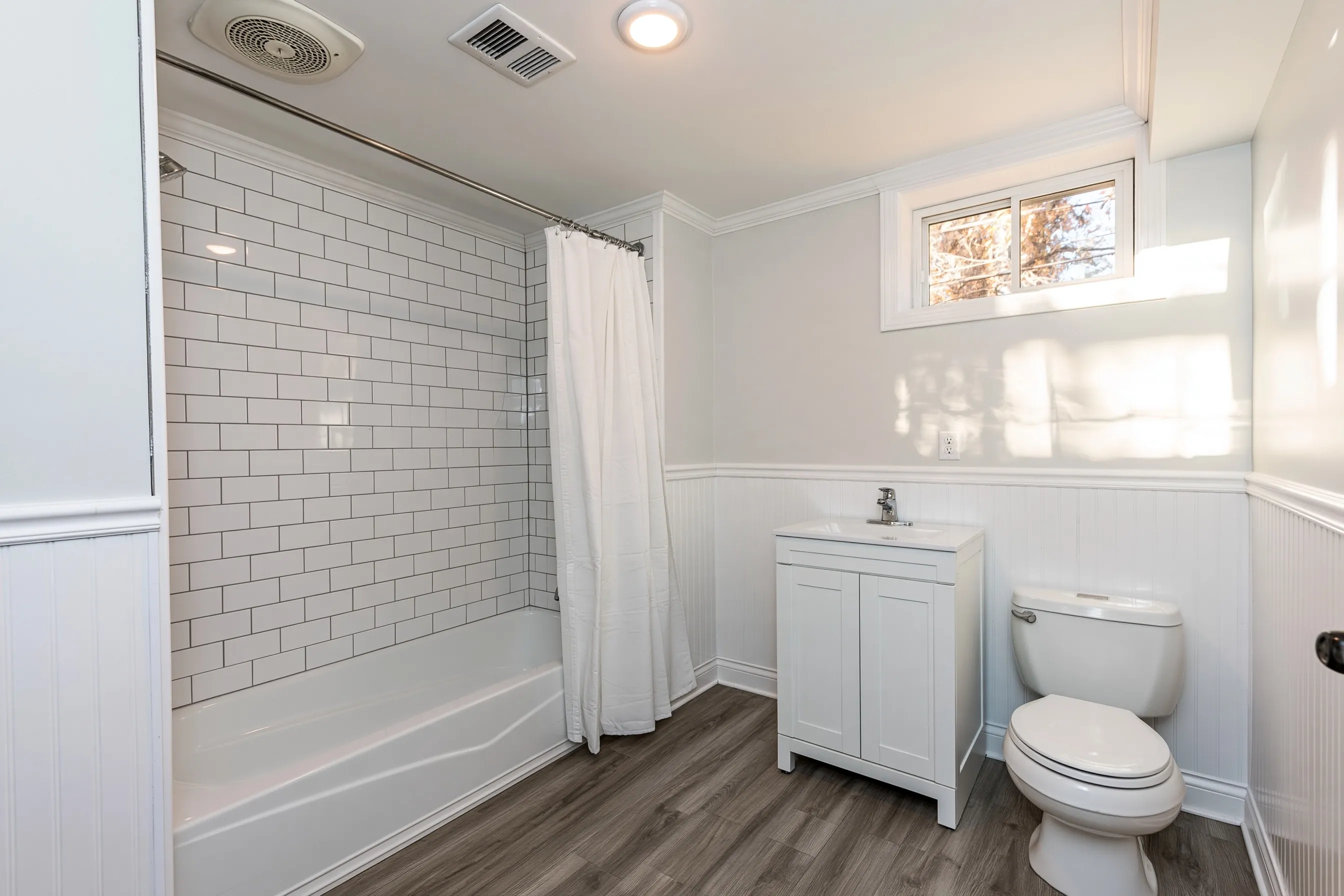 Basement bathroom with a tub and shower under white subway tile, a shower curtain, a white vanity, a toilet, white beadboard and a small high window