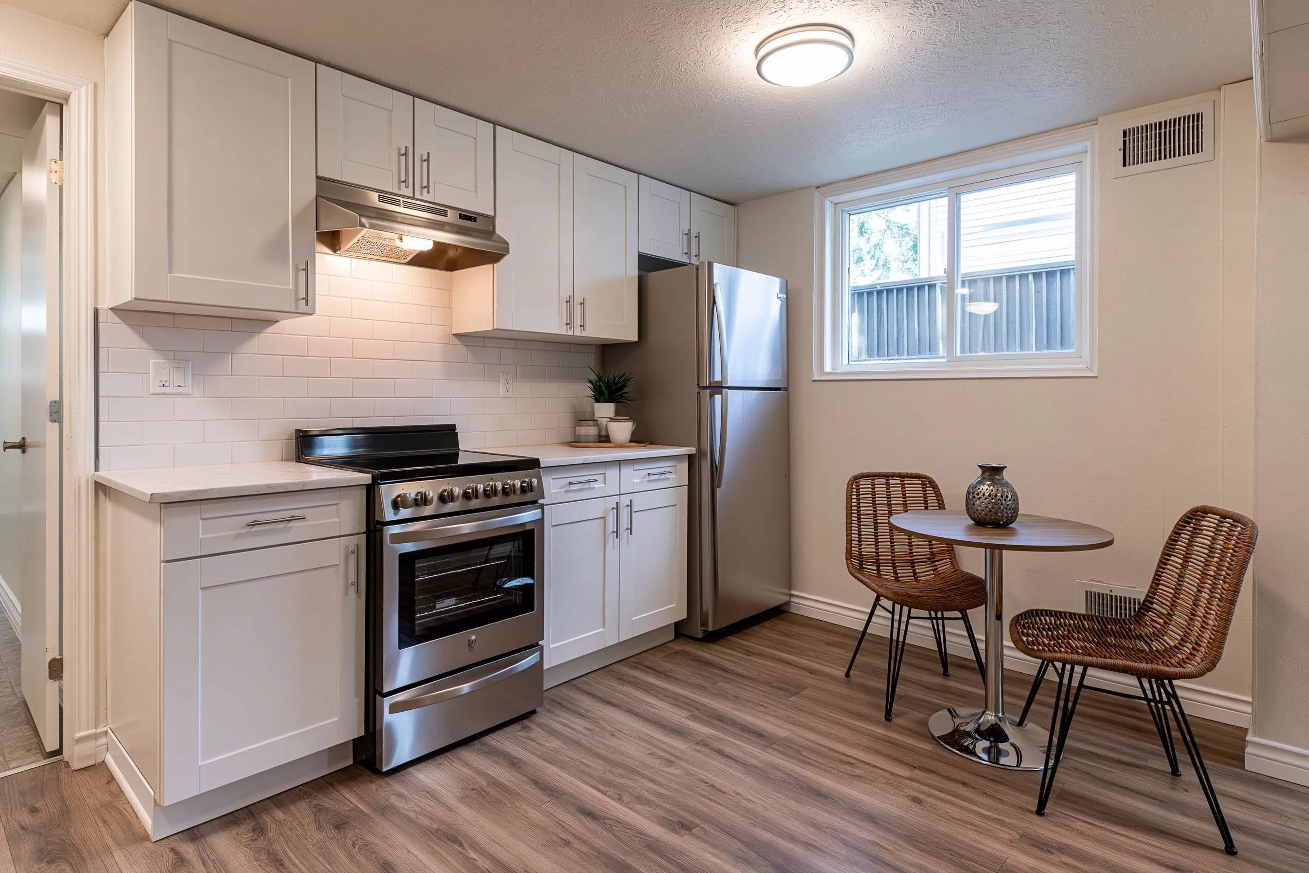 Finished basement kitchen with white cabinets, a range under a vent hood, a full-size refrigerator, a small dining table and a window beside it