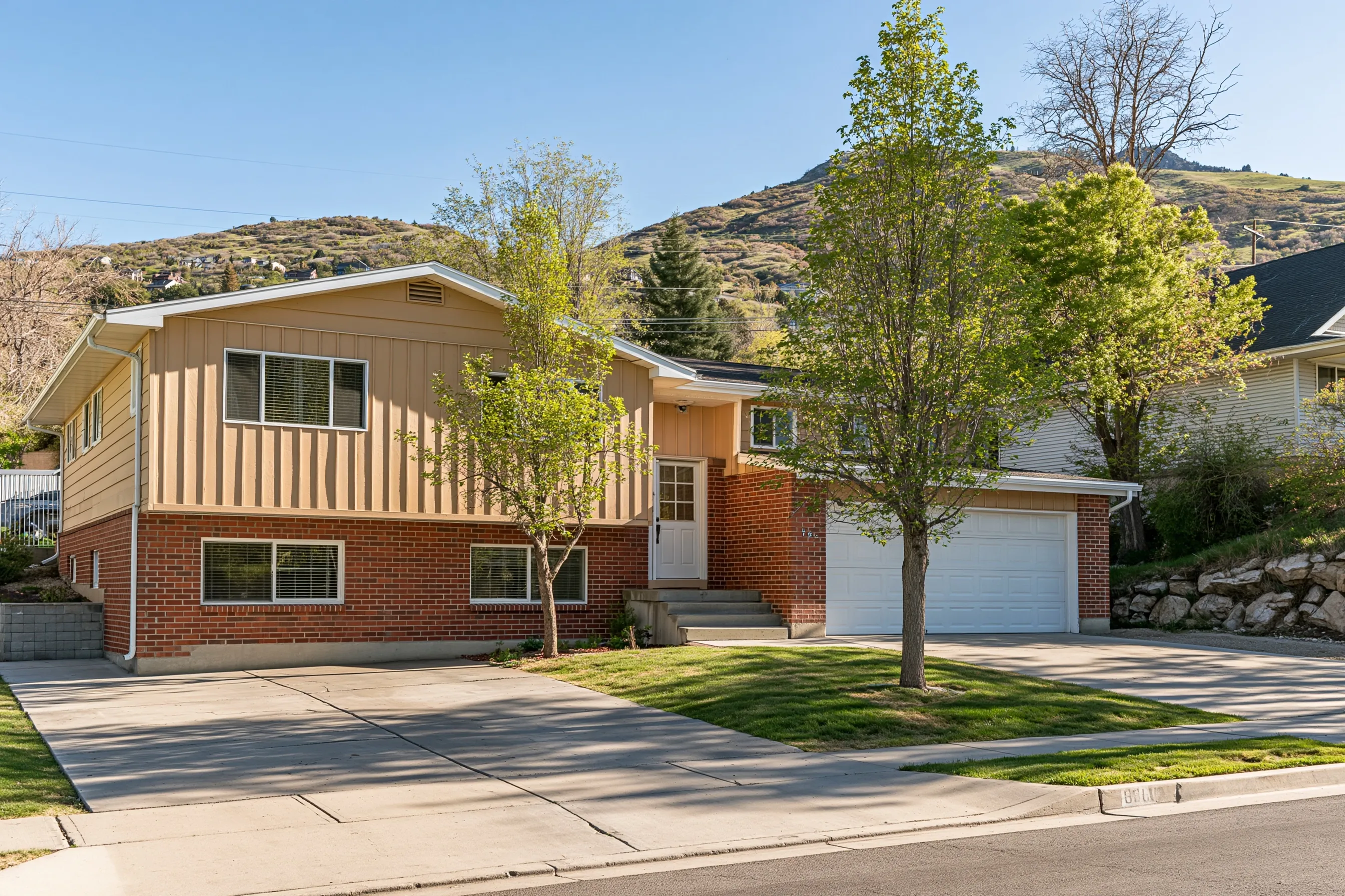 Front of a 1970s split-entry house with a brick lower level and light siding above, basement windows half above grade, a two-car garage and the Wasatch foothills behind